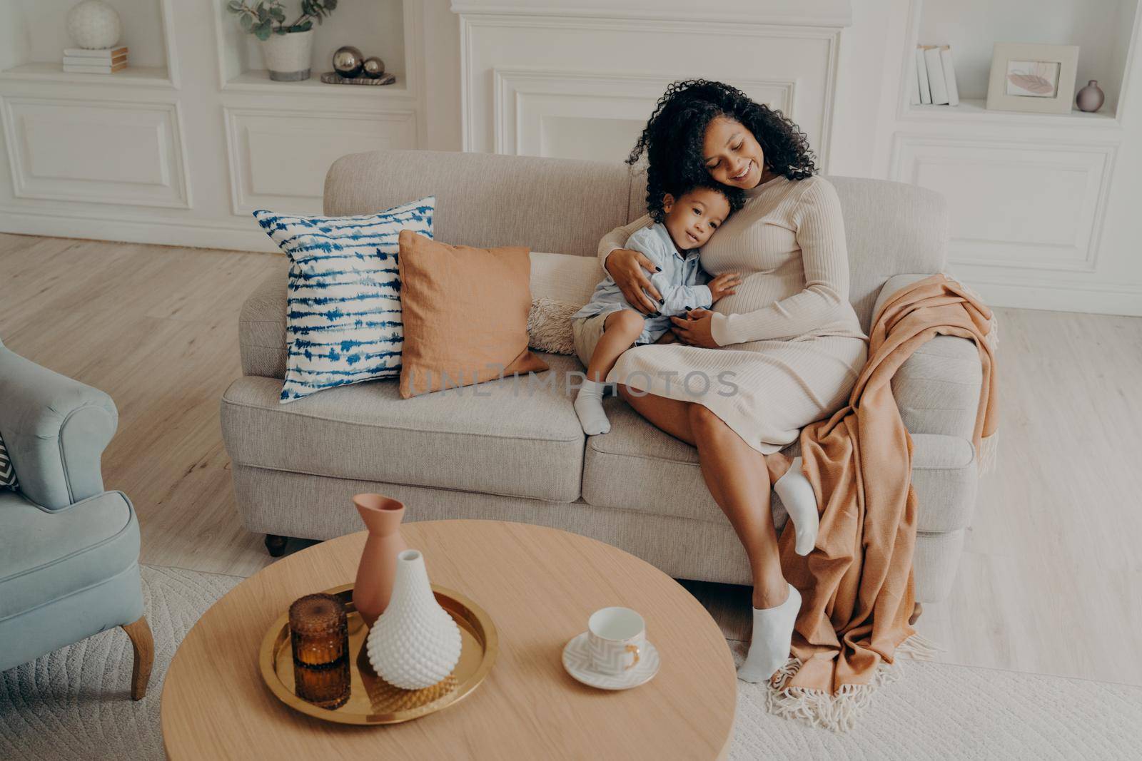 Single african pregnant mom tenderly embracing little son while sitting on cozy couch by vkstock