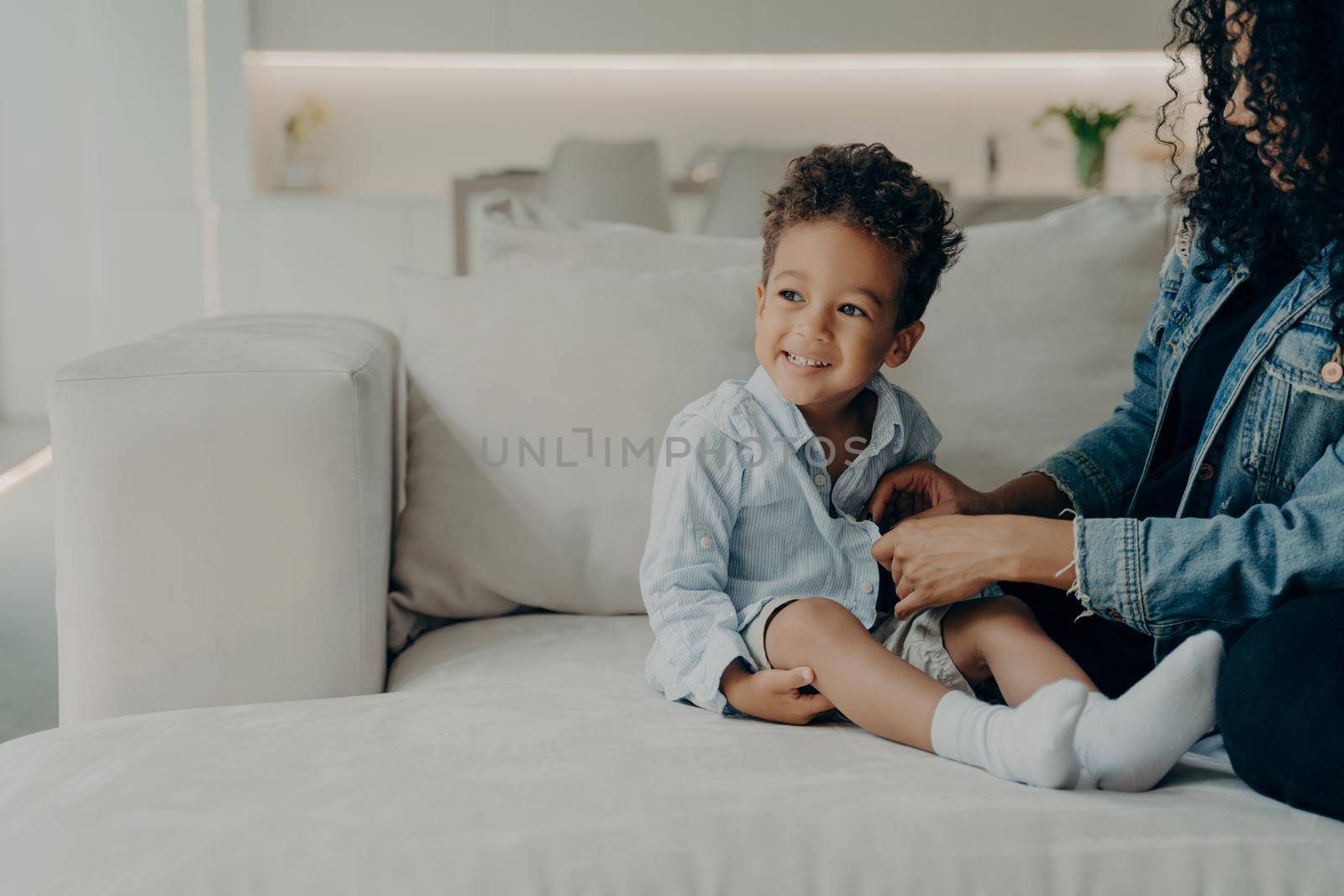 Young caring mother dressing up her little cute son with curly hair, putting on adorable boy shirt and buttoning it, getting ready to walk in park while sitting on sofa at home. Family and childcare