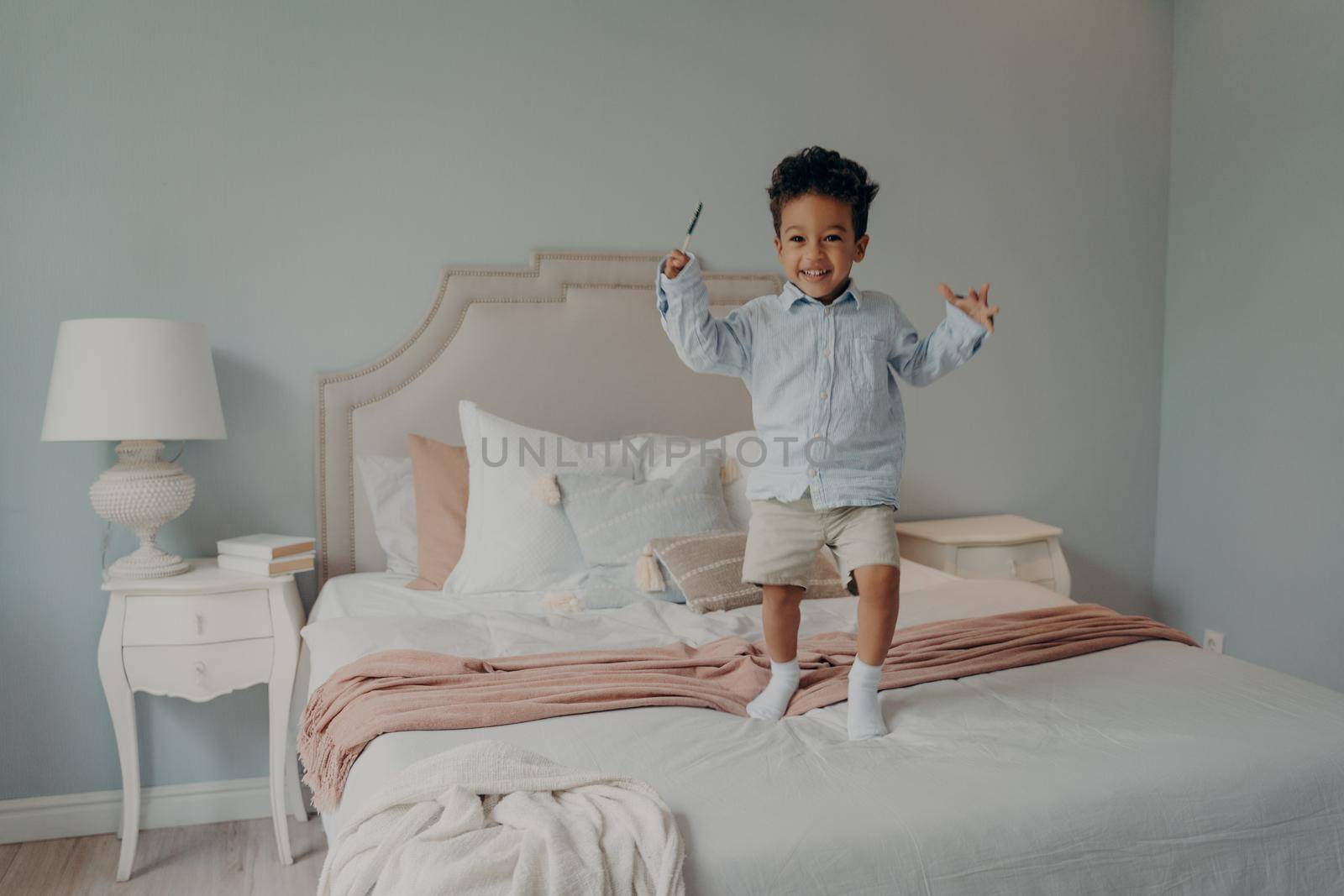 Joyful small afro american kid jumping on bed at home and smiling by vkstock
