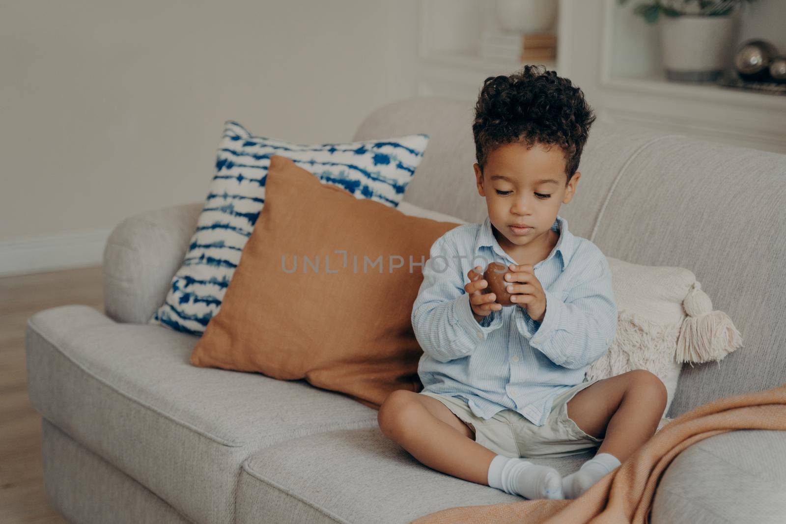 Adorable mulatto baby boy in casual clothes sitting on sofa decorated with pillows, holding chocolate egg and looking with interest expression on his face. Children and sweets concept