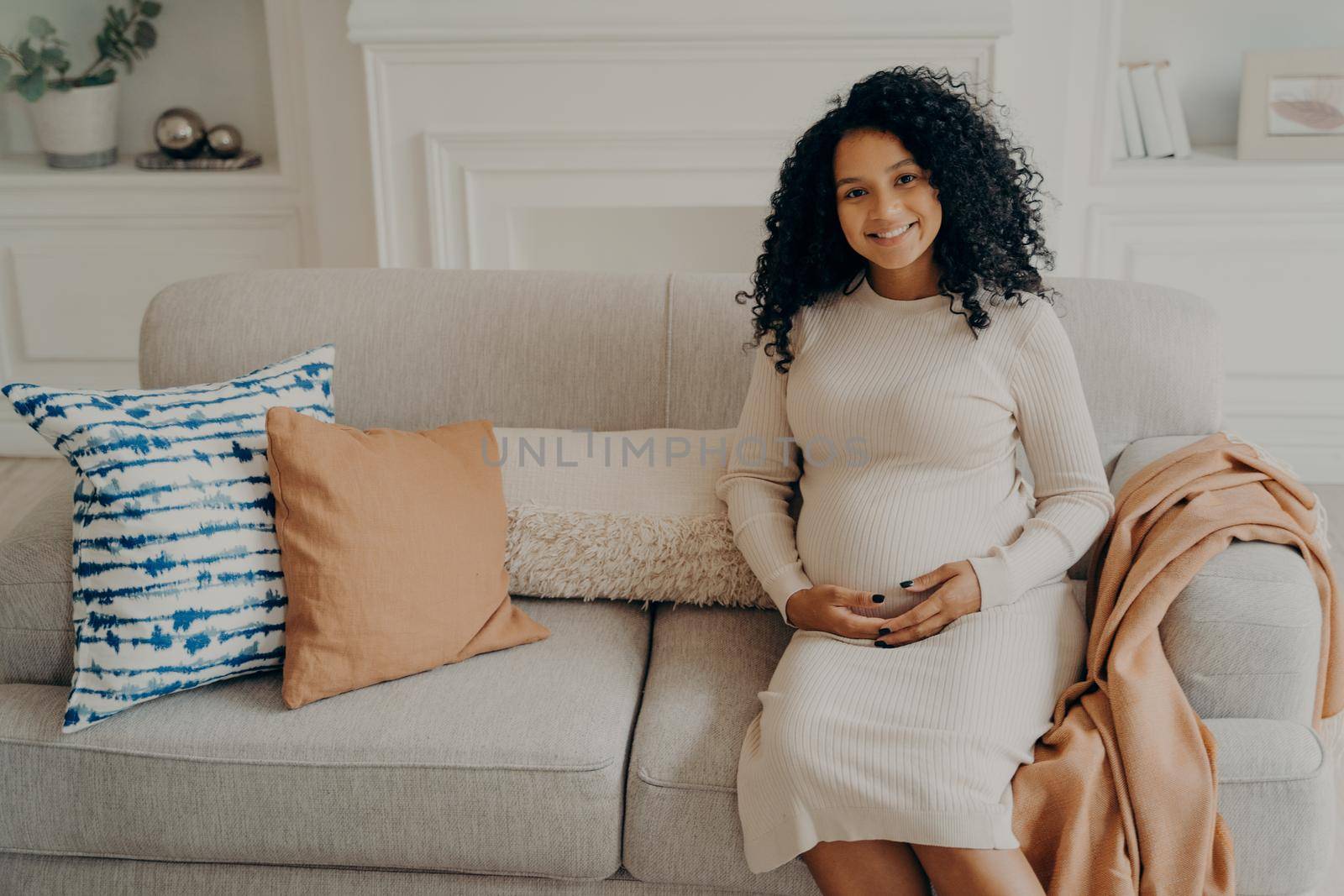 Beautiful african pregnant young woman sitting on sofa decorated with cushions, feeling happiness and love, keeping hands crossed on her bump belly in living room ambience, relaxing at home