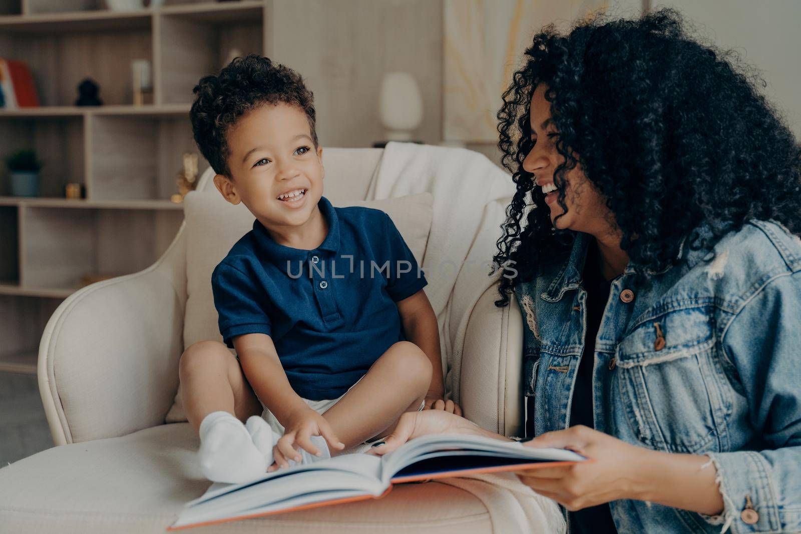 Passionate positive afro american woman mother reading funny stories to her cute excited little son, laughing together, spending time learning about animals, motherhood and child developing concept