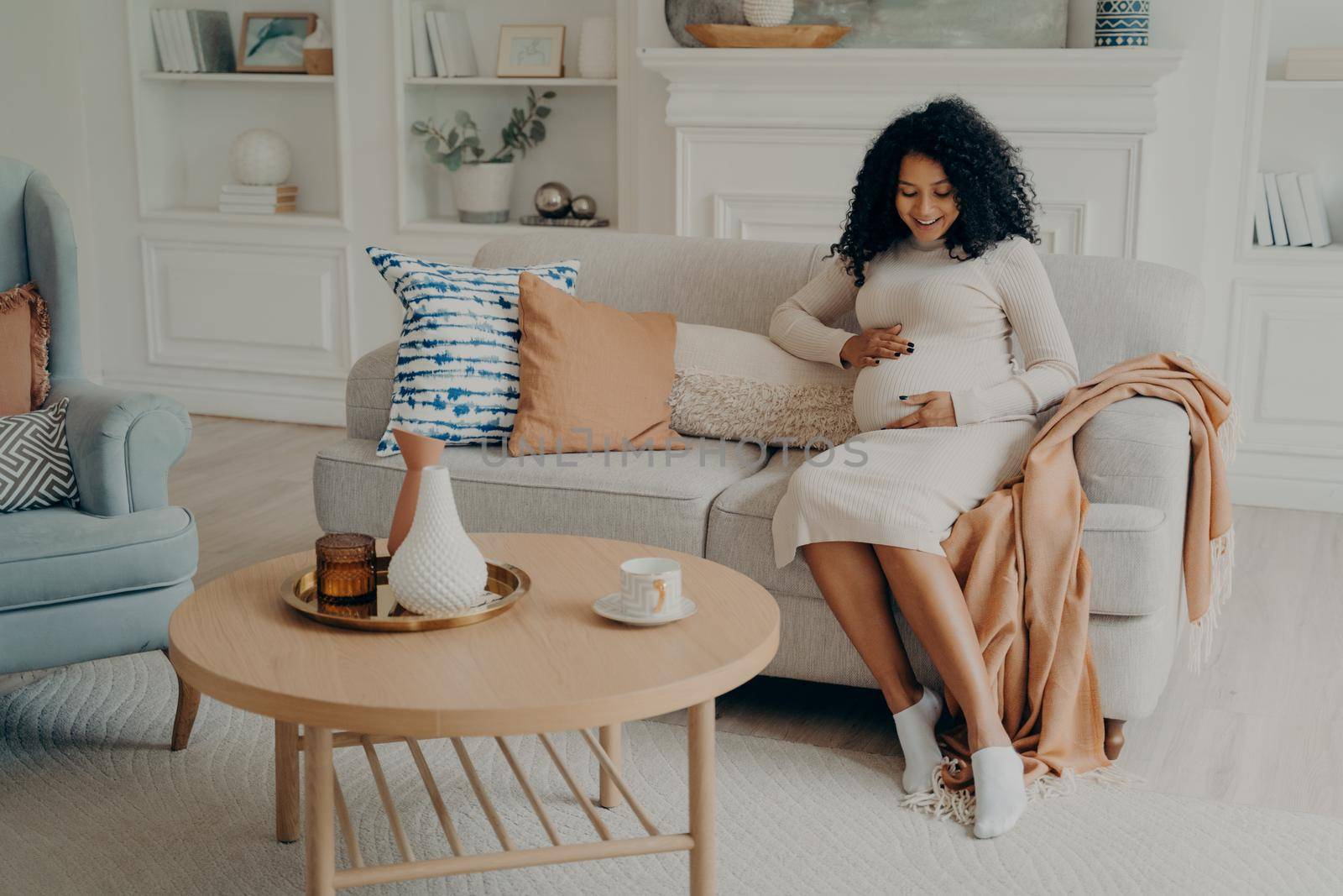 Beautiful pregnant mixed race woman in white dress with curly hair sitting and relaxing on couch in light themed living room at home, holding her belly and happy to feel baby move inside