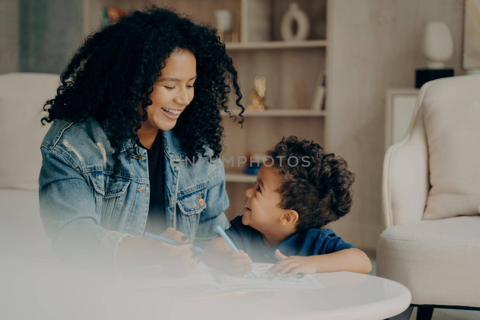 Portrait of happy joyful mixed race woman mom with long black curly hair sitting with little son in living room enjoying leisure time in each others company, learning how to draw. Happy maternity