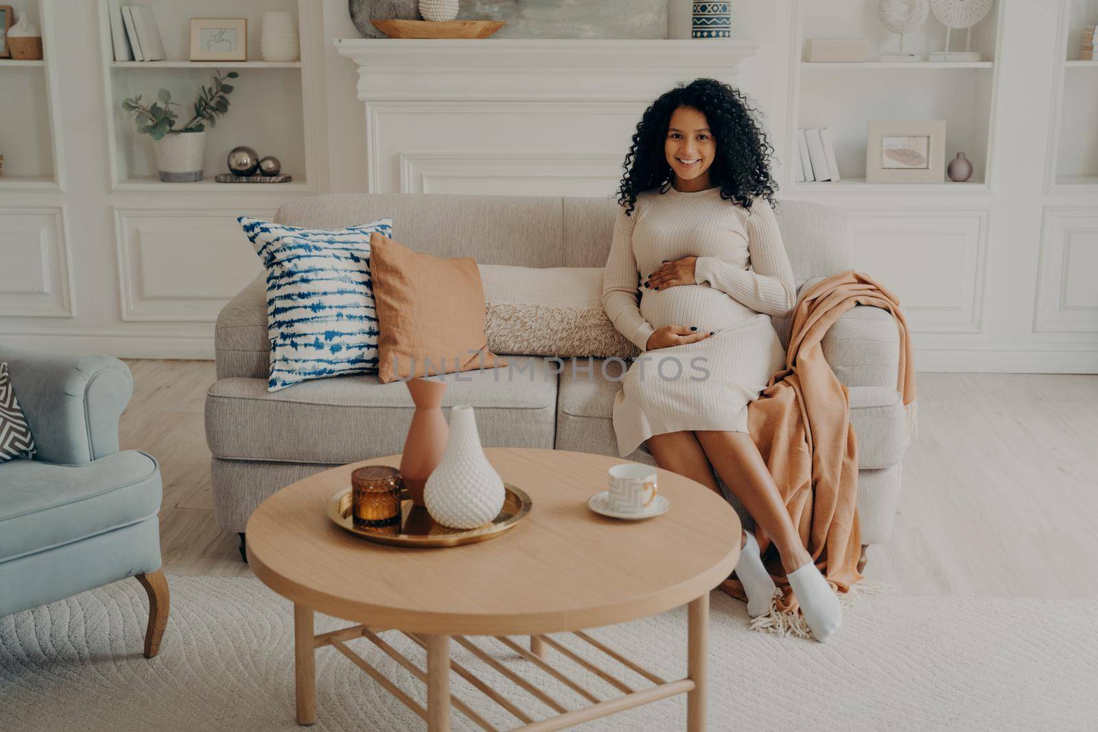 Charming afro american pregnant woman sitting on sofa with hands on her belly, expecting child, future mom looking joyfully at camera while relaxing living room at home, wearing knitted dress