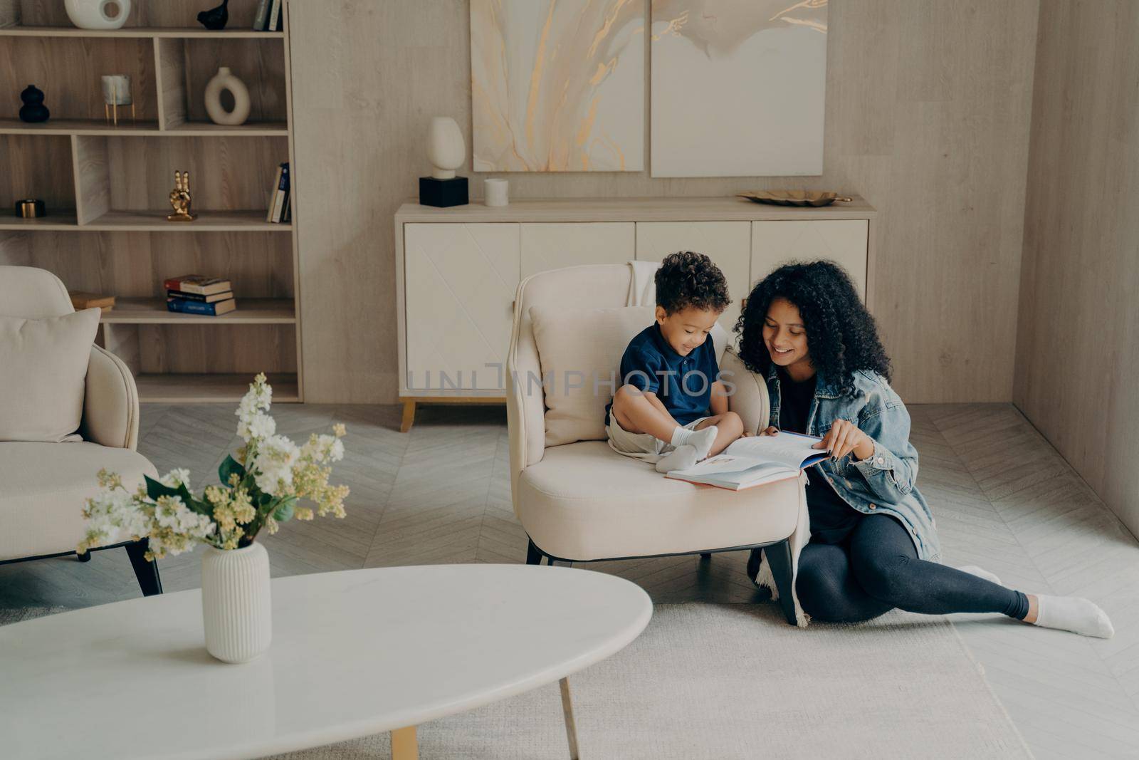 Little mixed race boy reading with his loving mom while enjoying time togerher in living room at home by vkstock