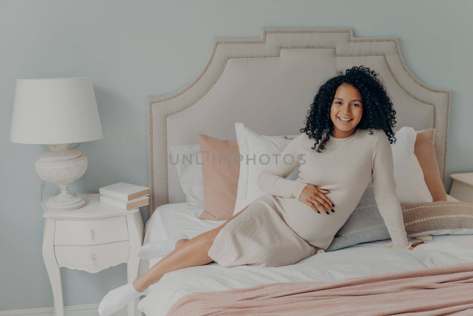 Healthy pregnant african american woman with curly hair relaxing on bed at home while being on third trimester, dressed in white dress looking happily at camera and getting ready for motherhood