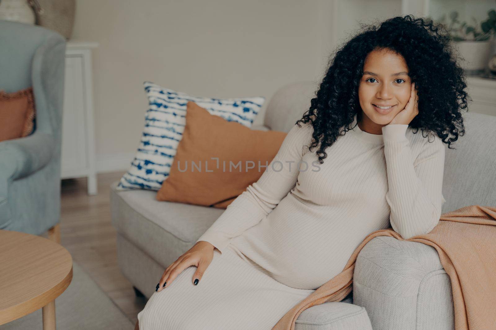 Beautiful happy african expectant mother sitting in relaxed pose while being pregnant, resting head on hand leaning on side of cozy sofa in living room, smiling and enjoying her leisure time at home
