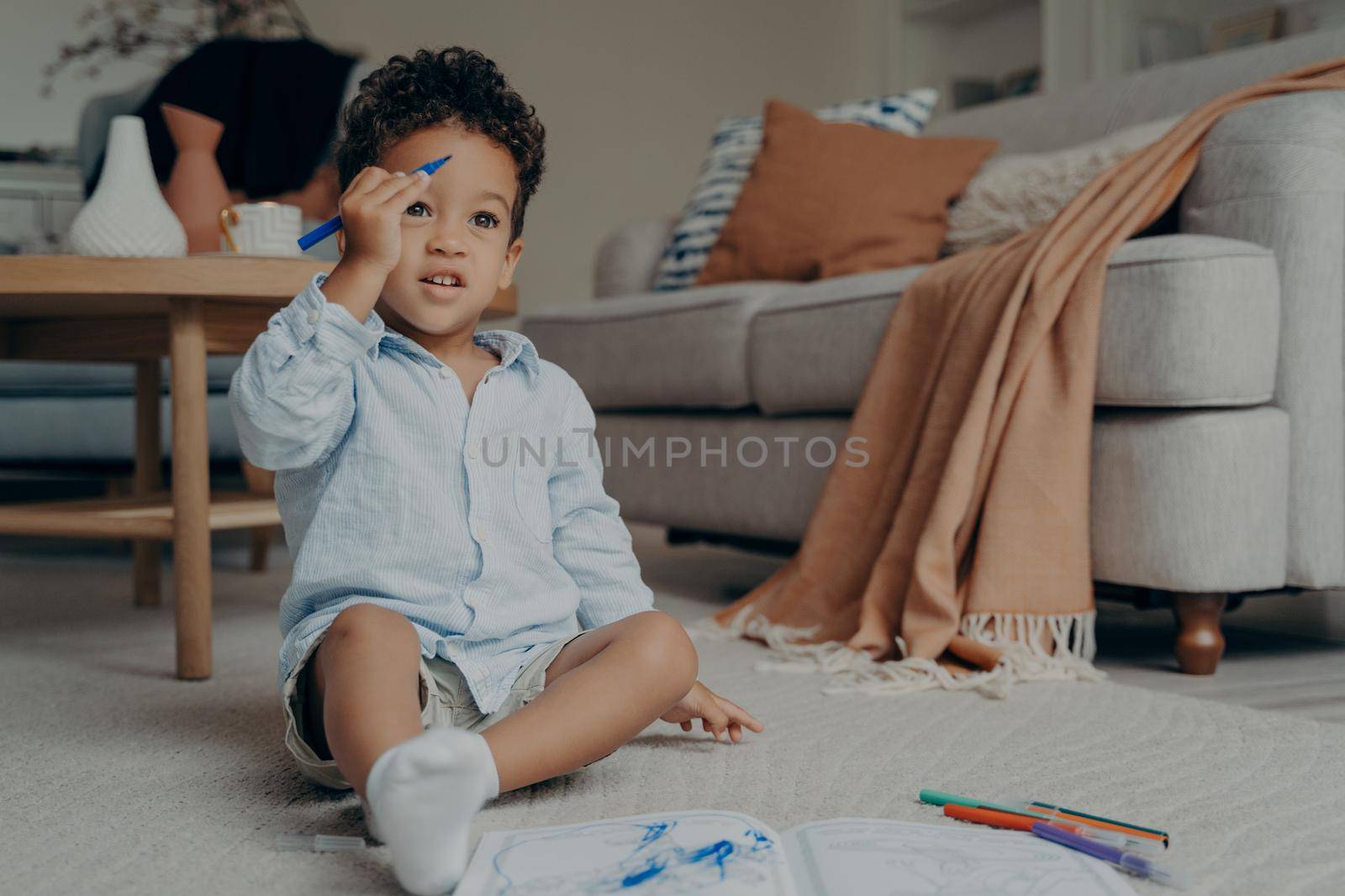 Mixed race toddler in casual clothes sitting on floor next to coffee table and cozy sofa in modern apartment, raised his hand up with blue felt tip pen. Creative leisure time for children indoor