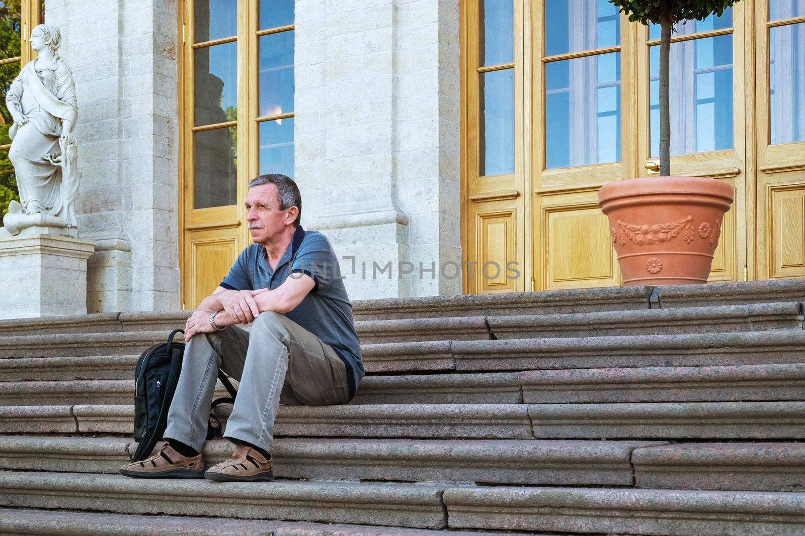 An adult caucasian pensioner in summer clothes sits and rests on the steps of an old historical house. Local tourism. Solo traveling. Selective focus.