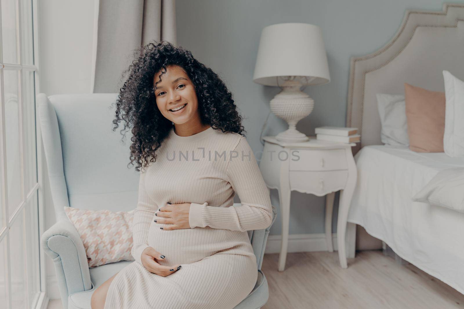 Happy pregnant afro american woman in white dress with curly hair, sitting on armchair in front of bedroom window, touching gently her belly and looking with smile at camera. Pregnancy concept