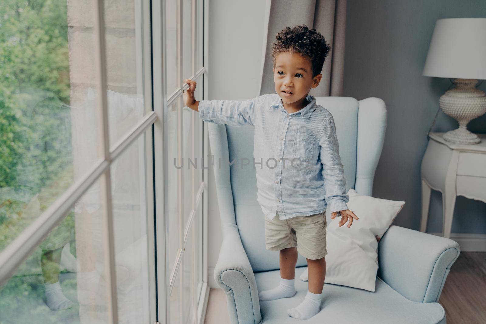 Full length of curious little afro american boy sweet toddler in stylish wear looking through window and waiting for mom while spending leisure time and playing alone at home in light room