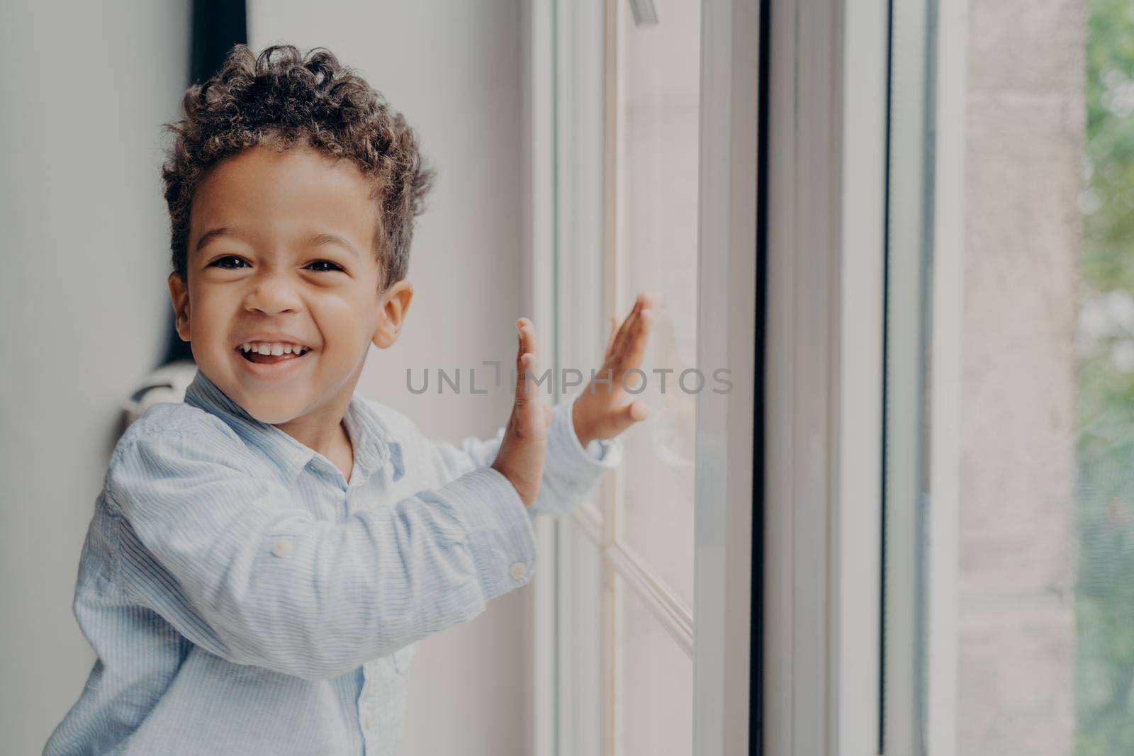 Portrait of little joyful kid with adorable smile and curly hair staying on windowsill next to closed large window, touching glass with small hands and smiling to camera in modern apartment interior