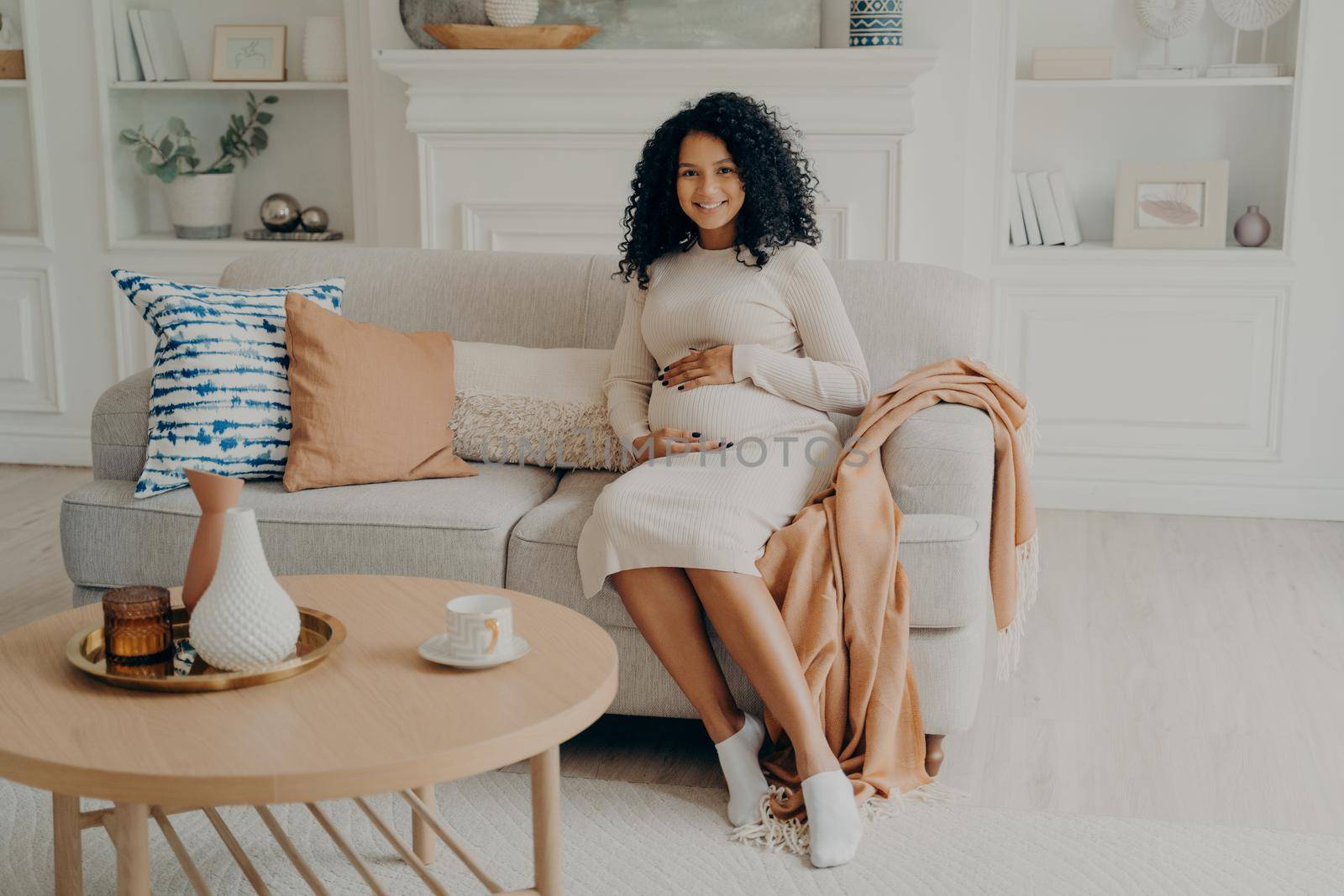 Afro American pregnant adorable lady in casual long dress sitting on couch decorated with cushions and blanket, holding her belly by hands and smiling on camera. Maternity and pregnancy concept