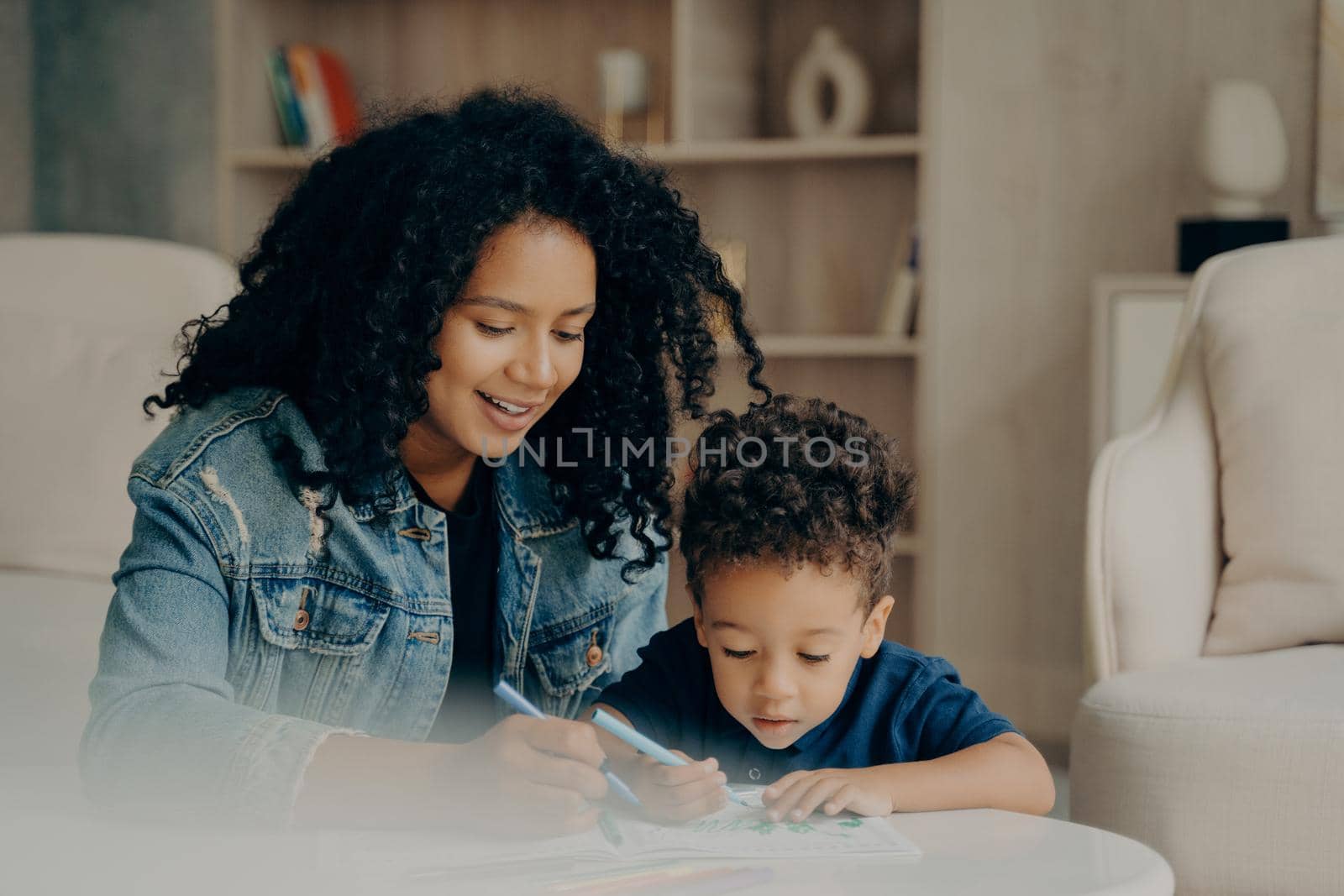 Pretty mixed race mom and little kid son focused on drawing with colored felt tip pens, playing in living room at home, spending leisure time together on weekend. Family and loving parents concept