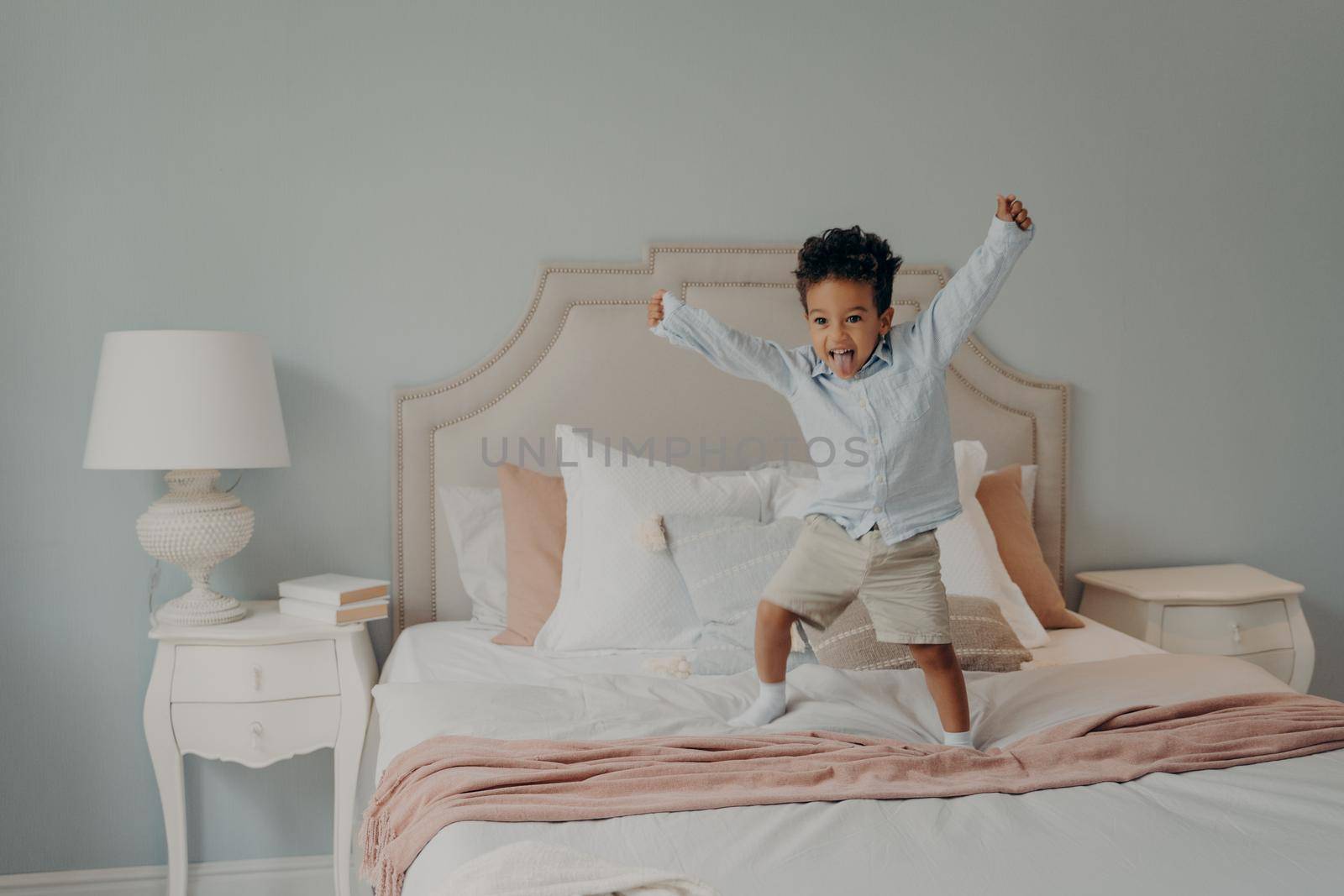 Adorable african cute kid jumping on bed covered with bedspreads in light pink and white tones next to lamp on bedside table, active mixed race child playing in parents bedroom during leisure time