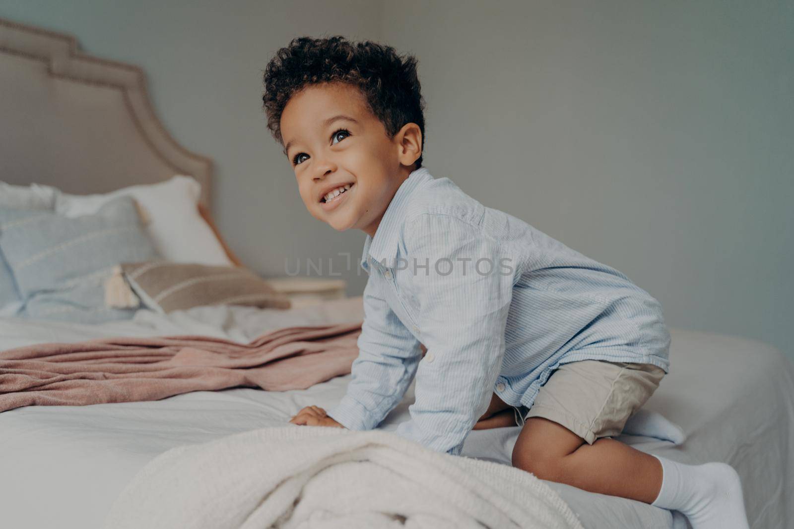 Smiling Afro American child trying to climb on big bed at home, having fun indoors by vkstock