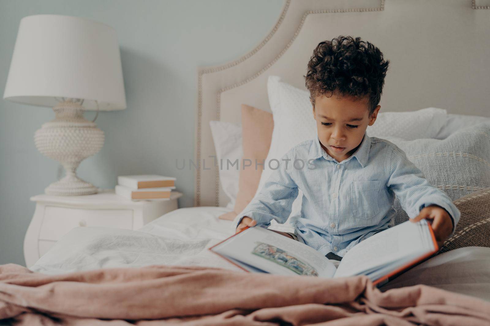Cute focused small boy in casual wear holds and tries to read childrens book, sitting on bed with nightstand next to with white elegant lamp on it. Kid spending weekend morning leisure at home