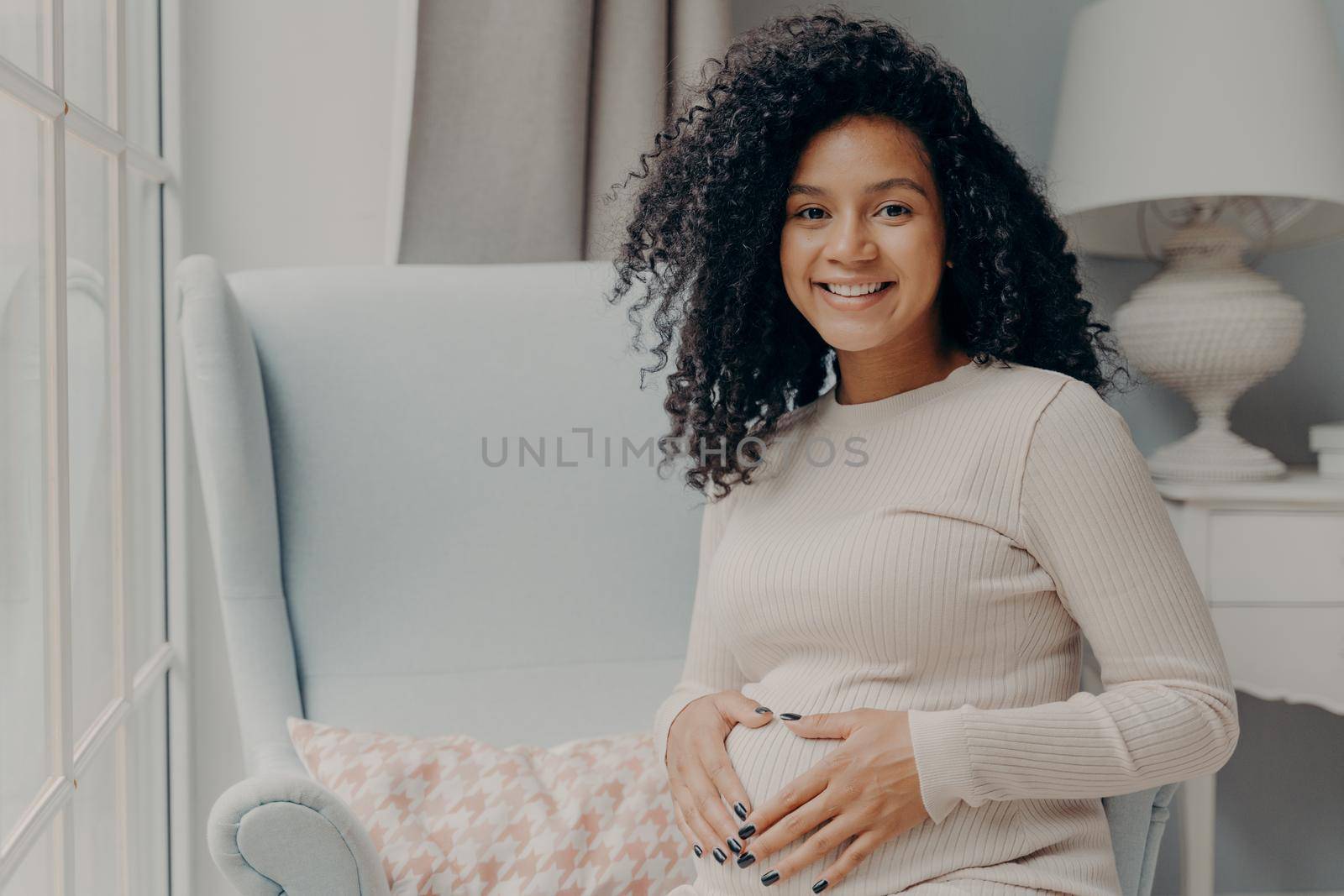 Portrait of young smiling pregnant african american lady in casual wear relaxing after walking outside on armchair in living room, keeping hands on belly and looking at camera with happy expression