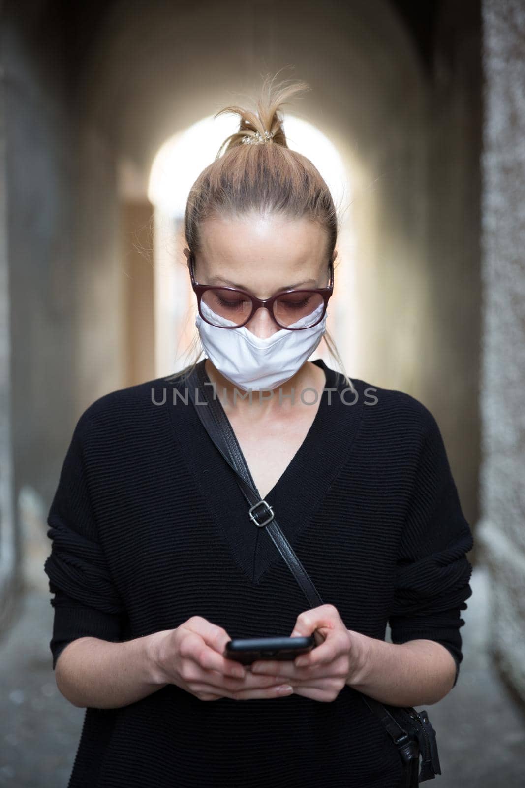 COVID-19 pandemic coronavirus. Casual caucasian woman at medieval city street using mobile phone, wearing protective face mask against spreading of coronavirus and disease transmission.