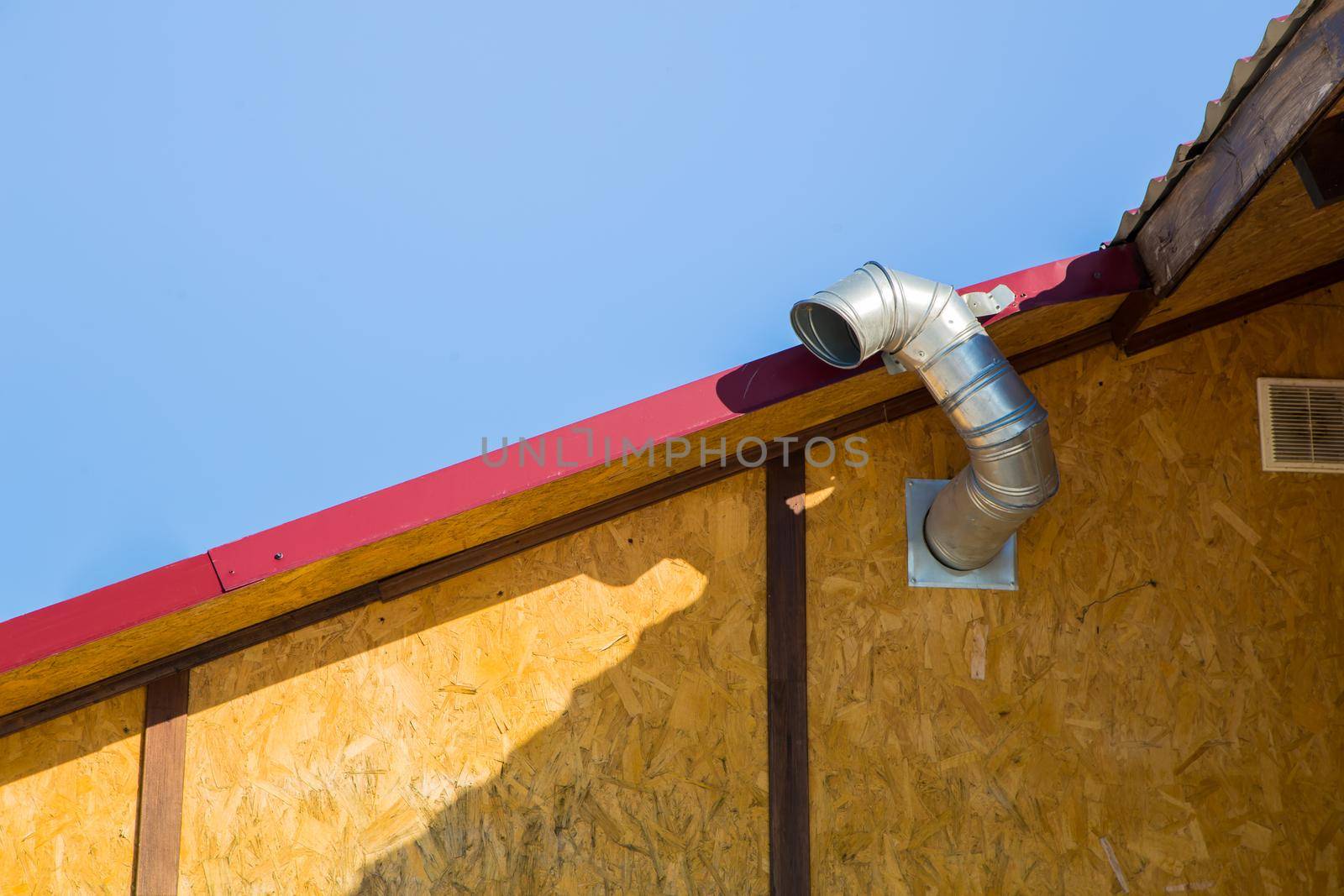 Chimney for exhausting smoke and polluted air in the wall against the blue sky. The bright morning sun illuminates the building and long shadows fall almost horizontally. Wooden house and red roof.