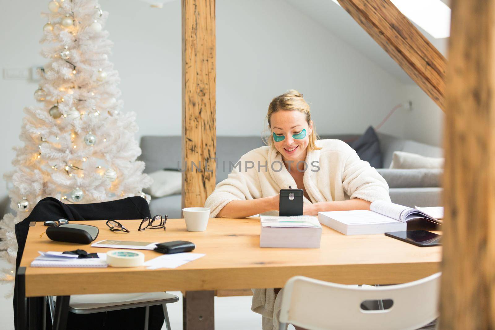 Young businesswoman wearing cosy warm bathrobe and cosmetic eye patches working remotly from home in winter Christmas time during 2020 corona virus pandemic. Work from home, selter in place, concept by kasto