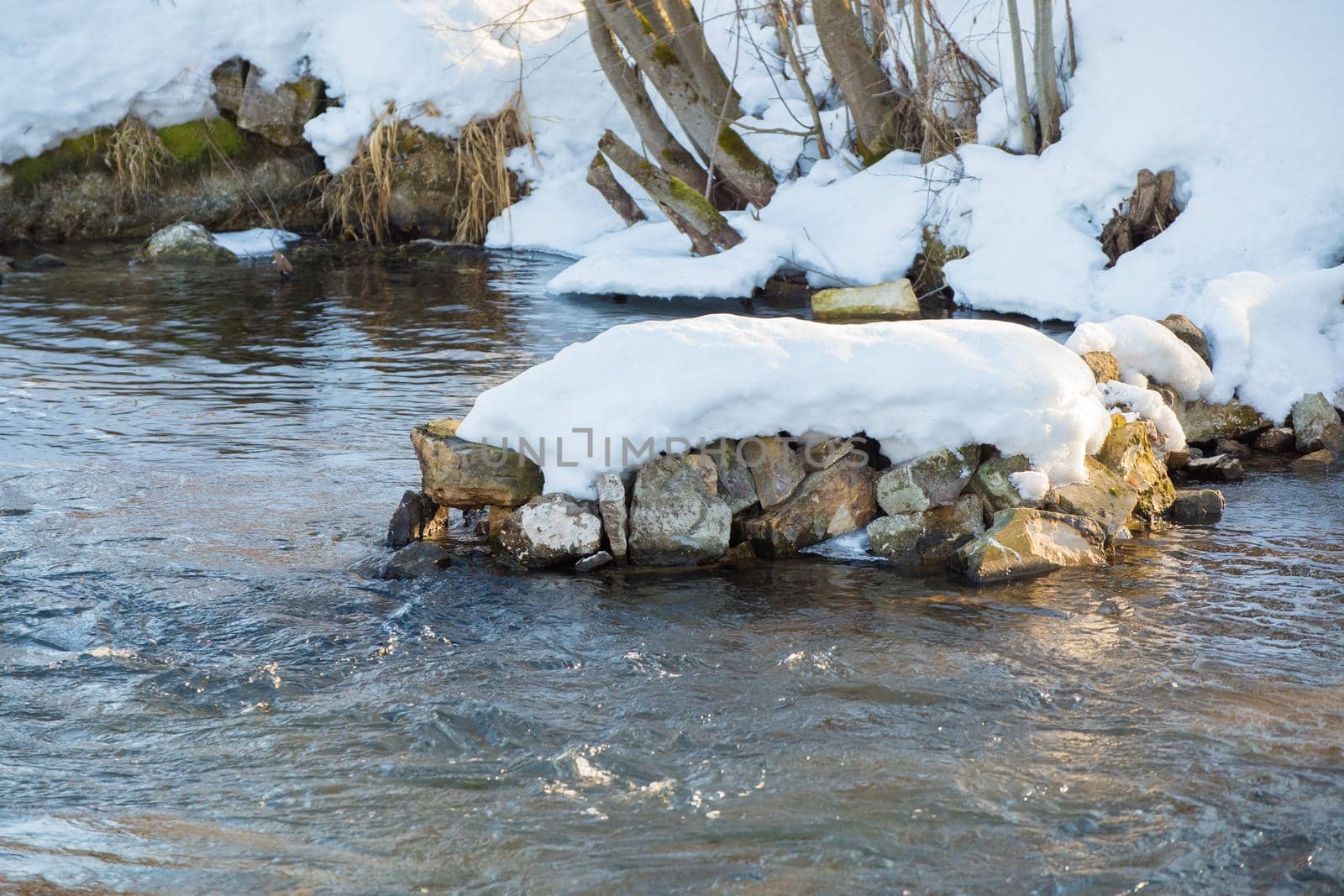 Winter landscape of a river with seething water and snow-covered banks. Snow lies on the grass and stones. A stormy stream of water with white foaming waves and ripples.