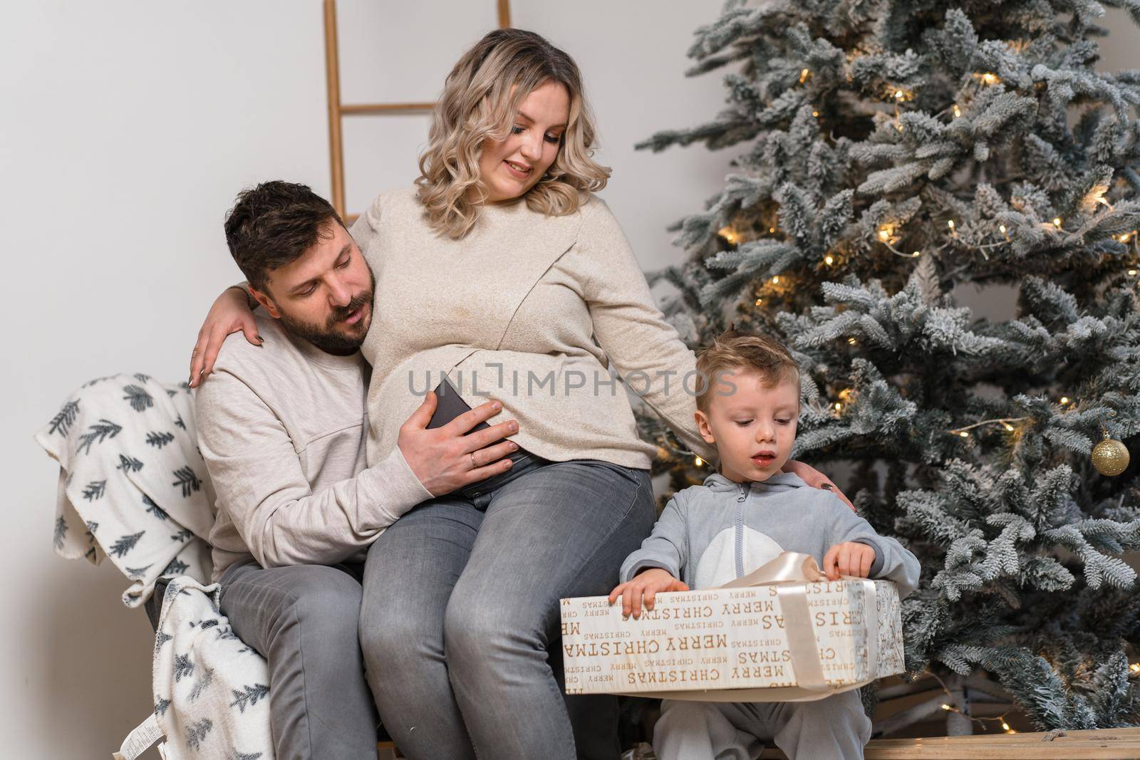 Christmas Family Happiness Portrait of dad, pregnant mom and little son sitting armchair at home near Christmas tree hug smile by andreonegin