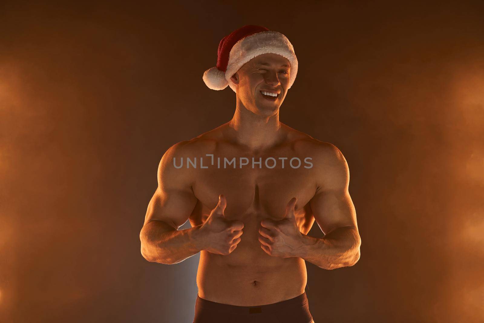 Portrait of muscular man wearing Christmas Santa hat, showing thumb up on smoky background by andreonegin