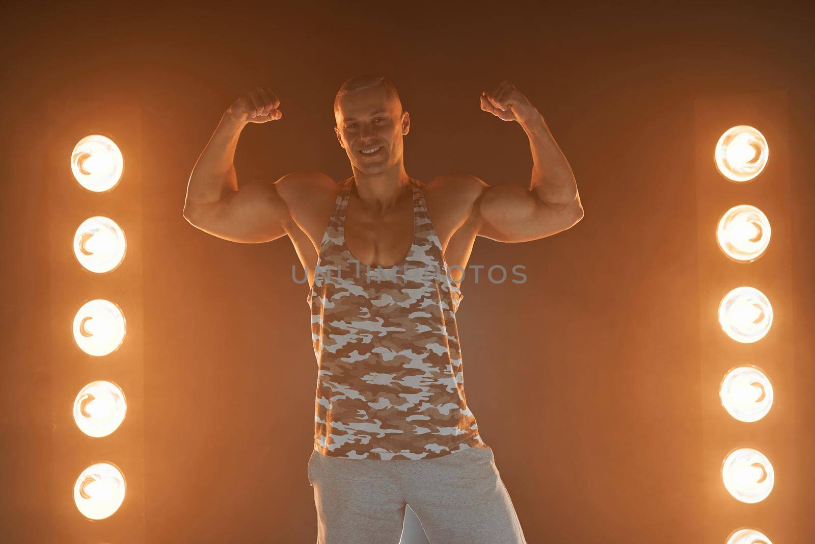 Active lifestyle. Professional bodybuilder showing biceps and smiling at camera, lamps illumination on background by andreonegin