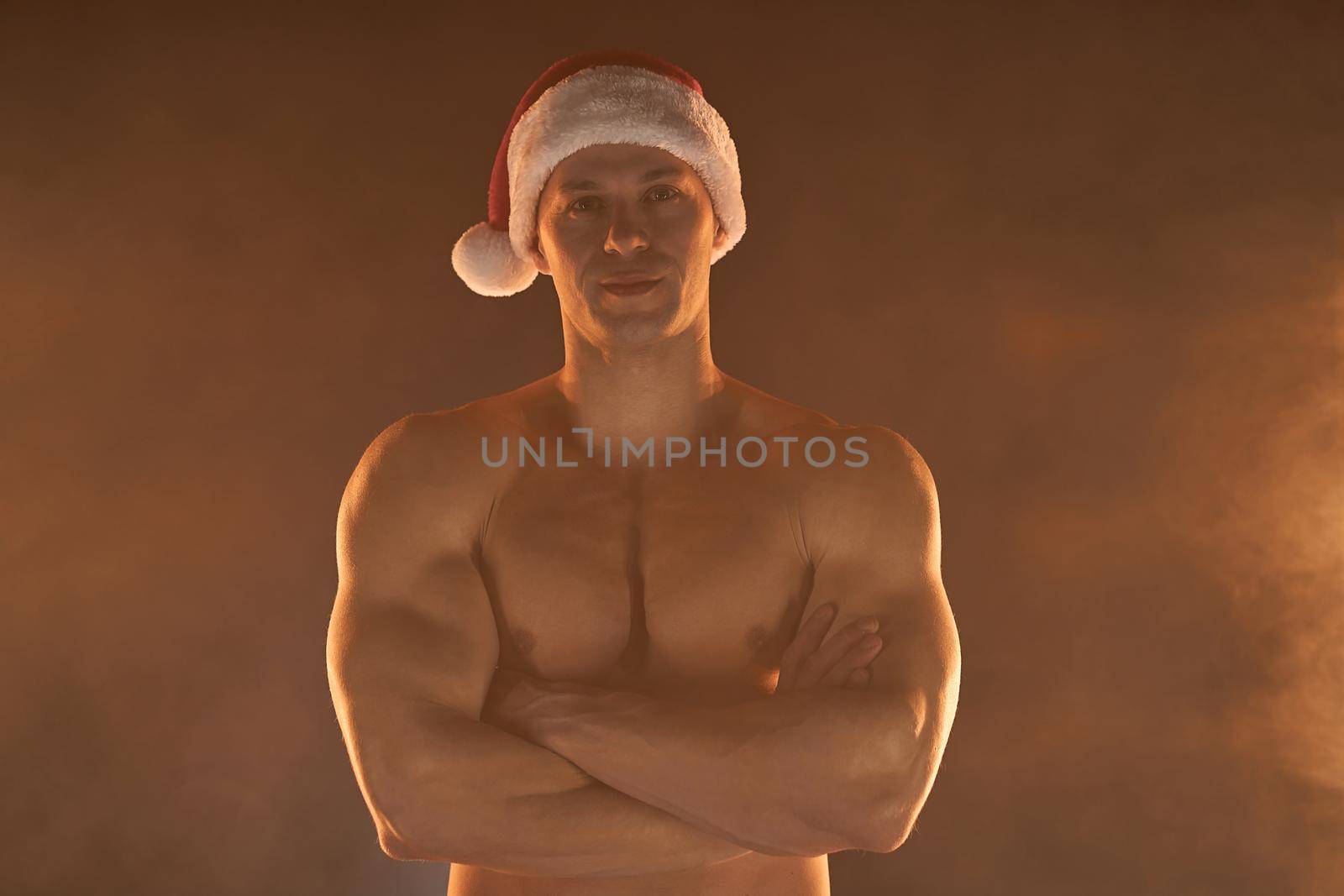 Portrait of muscular man wearing Christmas Santa hat, folded hands on smoky background by andreonegin