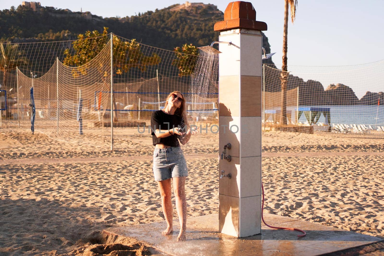 A girl in shorts and a black T-shirt on the beach near the shower. Shower on the beach. Nearby there are palm trees and a sports volleyball court.