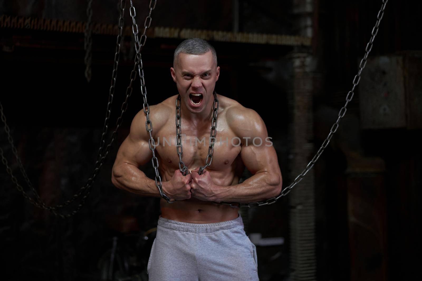 Primal instinct. Angry muscular man screaming at camera and breaking chains on chest, dark background