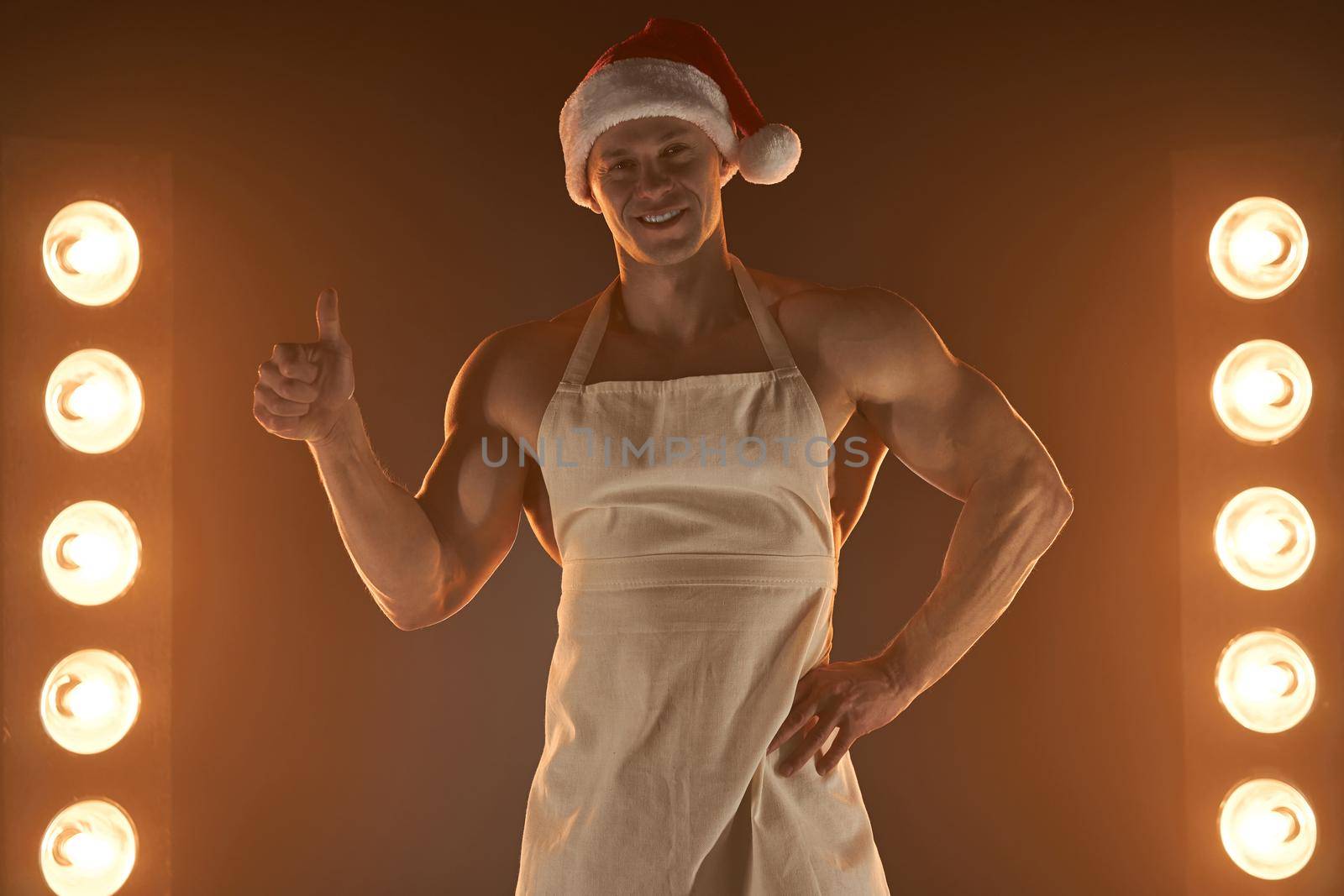 Hot Christmas dessert. Muscular man wearing apron and Santa hat showing thumb up gesture and smiling at camera, lamps illumination on background by andreonegin