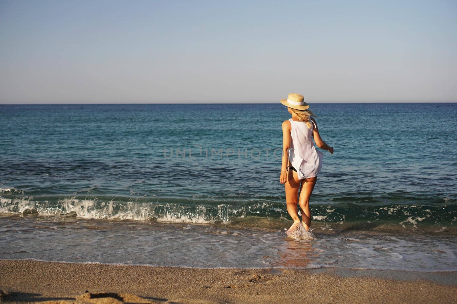 The girl walks on the beach. Beautiful blonde woman in white blouse and straw hat enjoying the sea