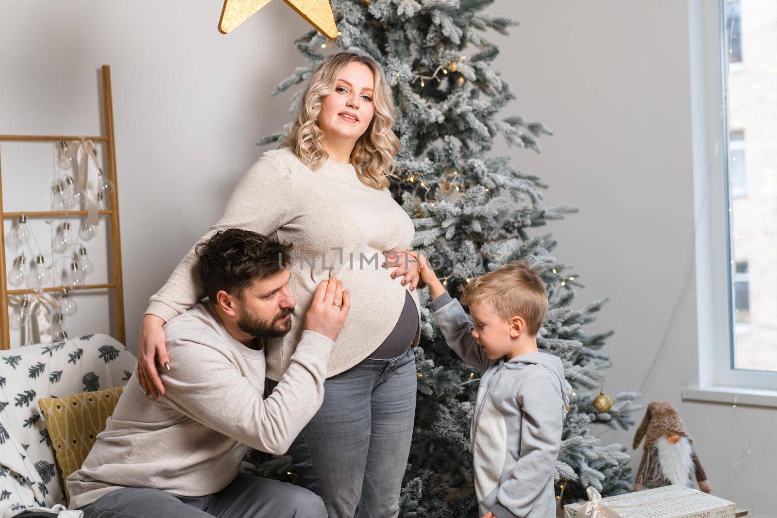 Christmas Family Happiness Portrait of dad, pregnant mom and little son sitting armchair at home near Christmas tree hug smile by andreonegin