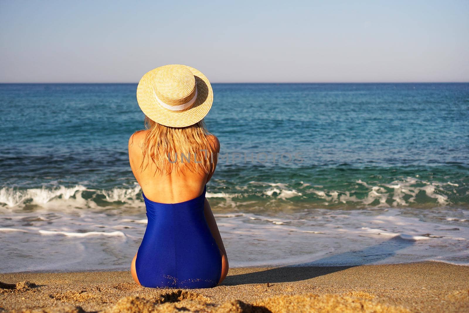 Young Woman Sunbathing At Tropical Beach. Woman in a blue swimsuit and hat sits on the seashore on sand