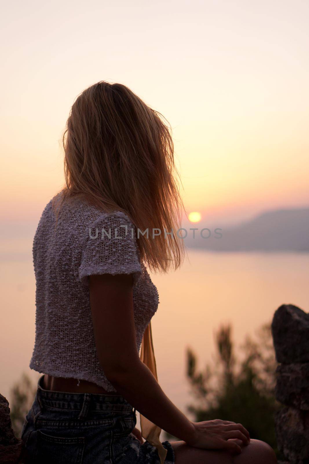 The girl looks at the sea sunset from a high point. Vertical photo with woman back