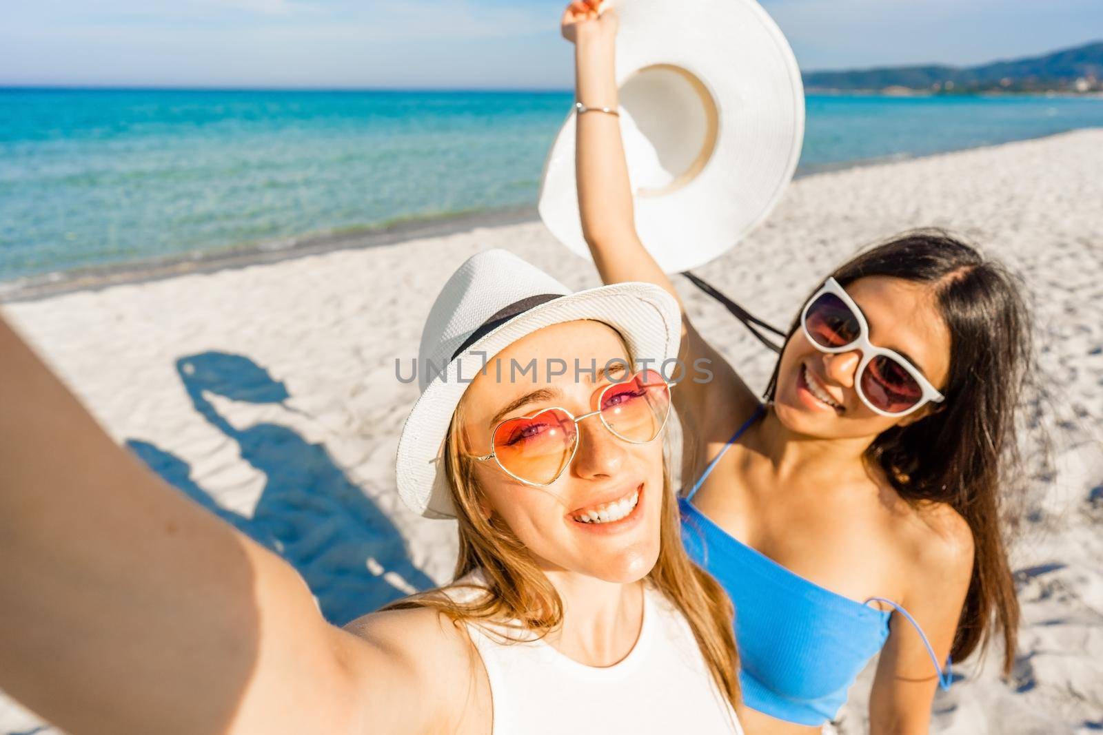 Two beautiful girls with white hats taking a self-portrait enjoying summer vacation on tropical beach resort. POV of young blonde woman using smartphone to photograph herself with best teen friend by robbyfontanesi