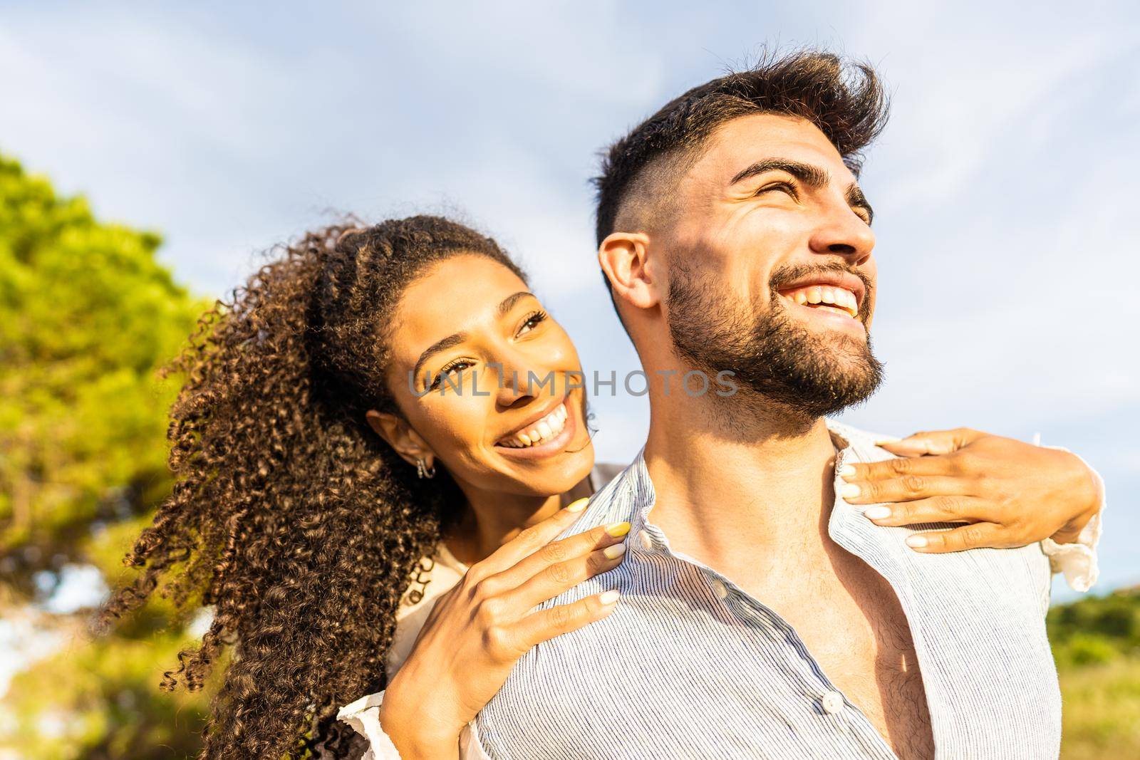 Multiracial happy young beautiful couple in love having fun outdoor in nature at dusk looking at horizon kissed by the setting sun. Toothy smiling Afro-American girl embracing from back her boyfriend by robbyfontanesi
