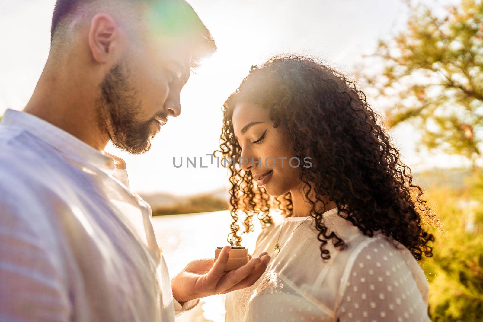 Backlit suggestive romance scene of young beautiful multiracial couple in love. Handsome guy making the marriage proposal to her black Hispanic girlfriend showing engagement ring close to her