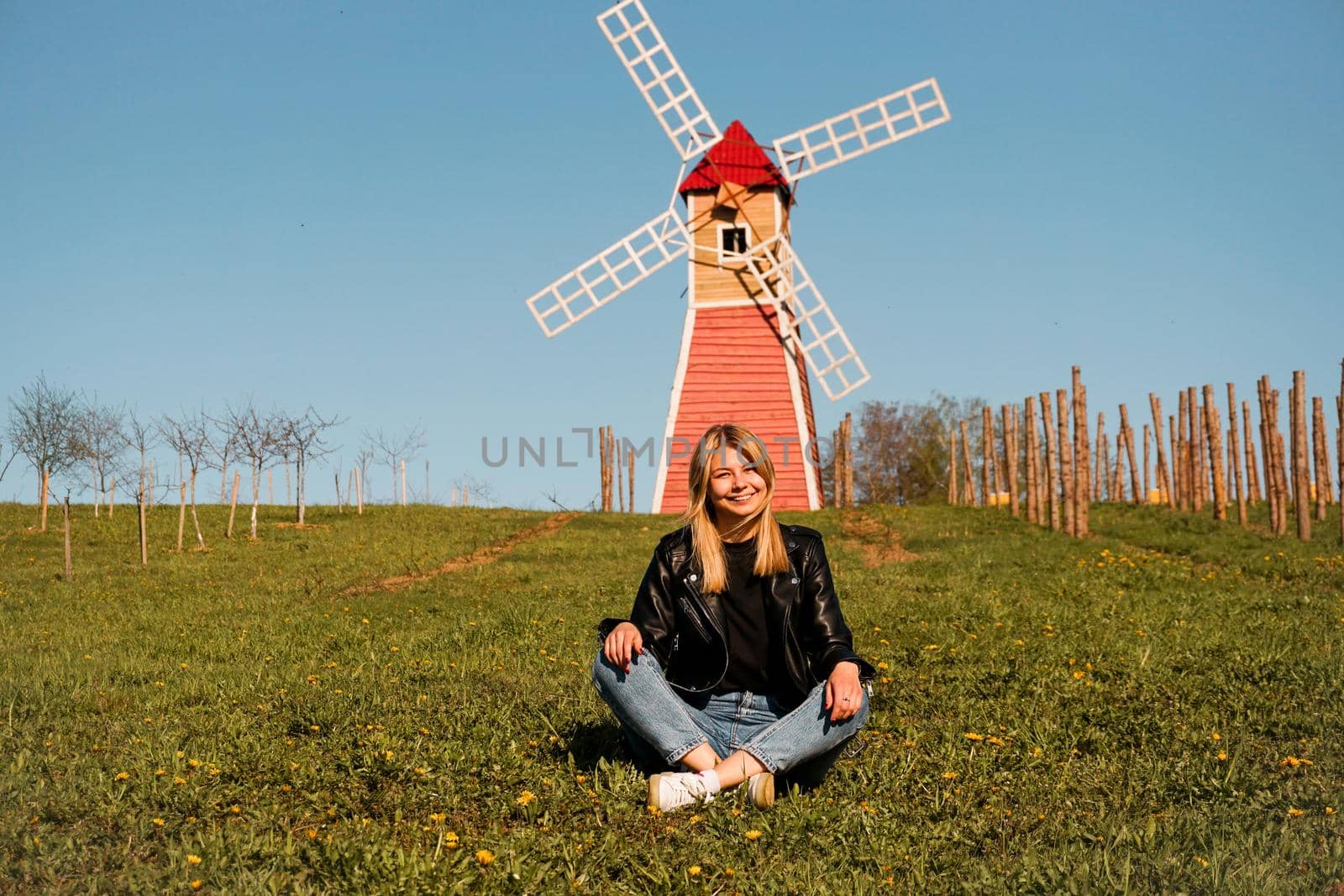 Beautiful young woman sits on the grass against the backdrop of the red mill. Sunny summer day. Rest in the countryside