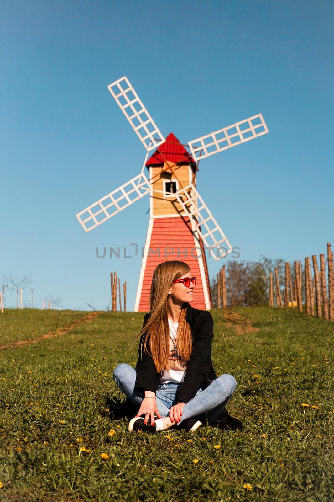 Beautiful young woman in red sunglasses sits on the grass against the backdrop of the red mill. Sunny summer day. Rest in the countryside