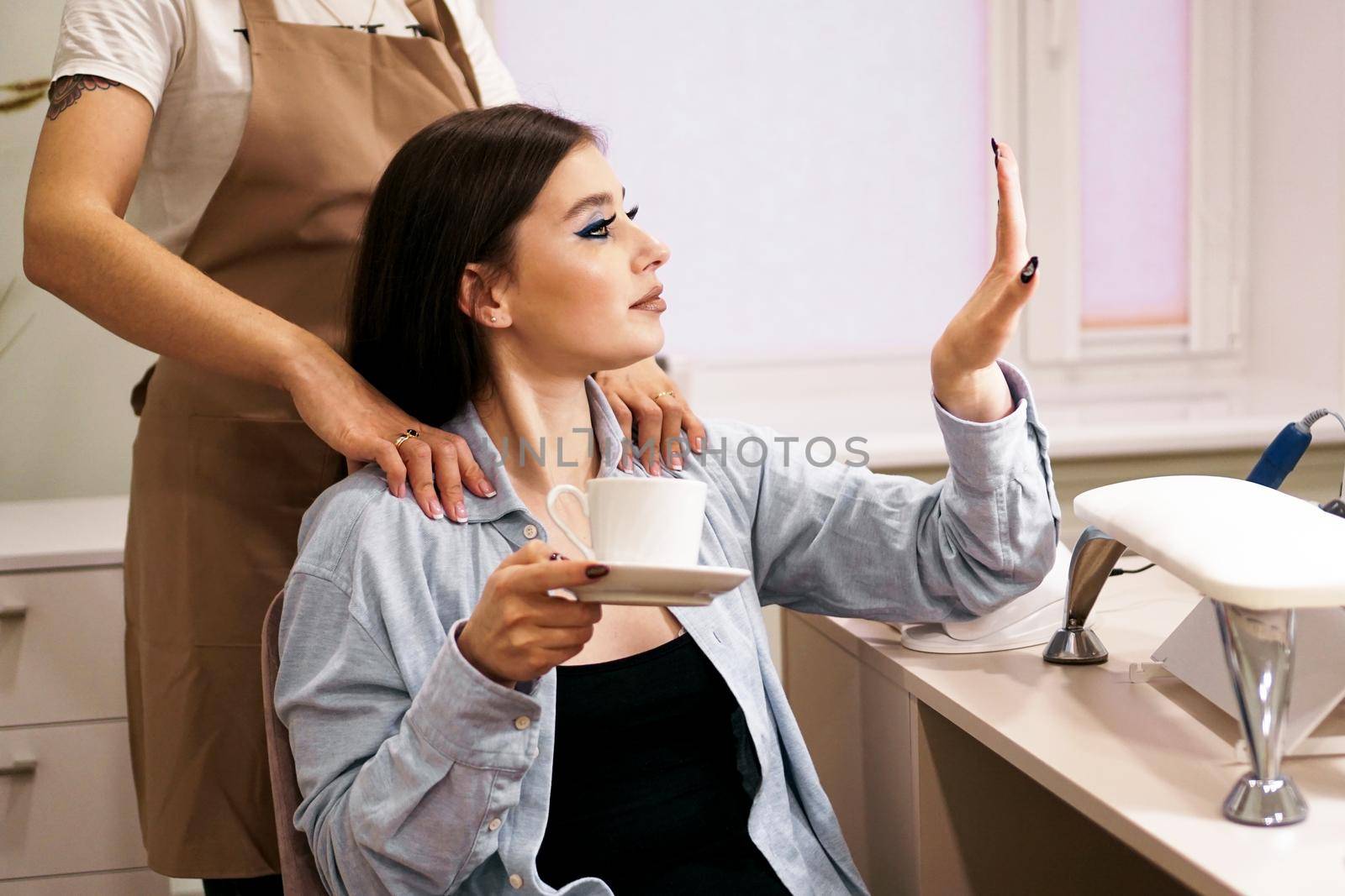 The girl looks at her manicure during the massage in beauty salon. She drinks coffee