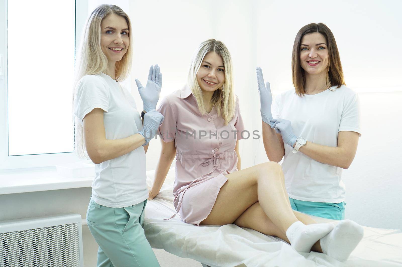 Two young beauticians or doctors put a glove on their hand and smile. The patient lies on the couch and smiles