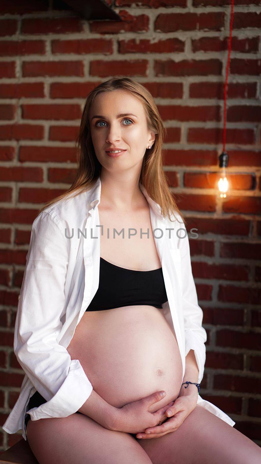 Beautiful young stylish pregnant woman, in black lingerie and white shirt, in the interior of a loft on the background of a brick wall