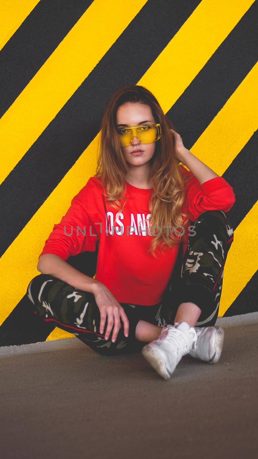 Fashion model wearing red hoodie with the inscription los angeles and big plastic glasses posing in the city at parking. Fashion urban outfit