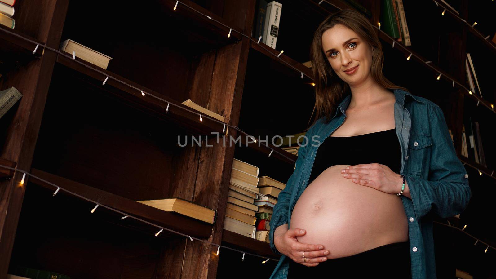 Beautiful young stylish pregnant woman, in black lingerie and blue shirt, in the interior of a loft on the background of wooden bookcase with books