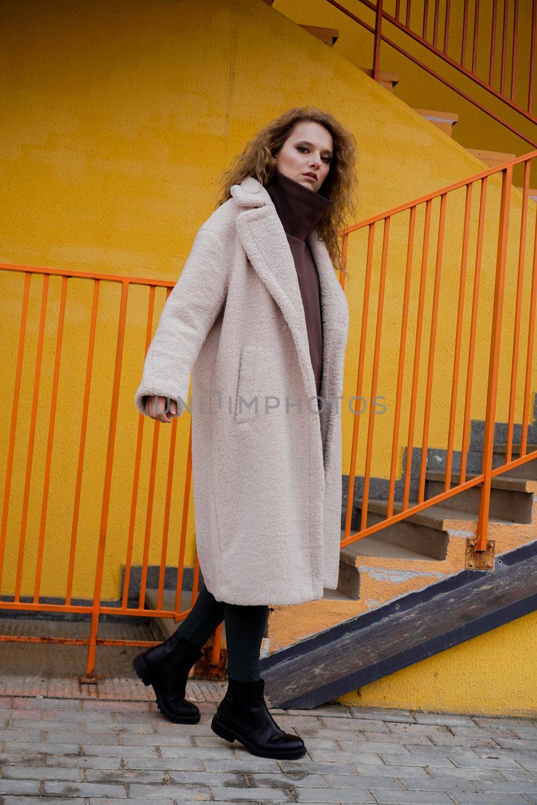 A girl with red curly hair in a white coat poses on the yellow orange parking stairs. City Style - Urban
