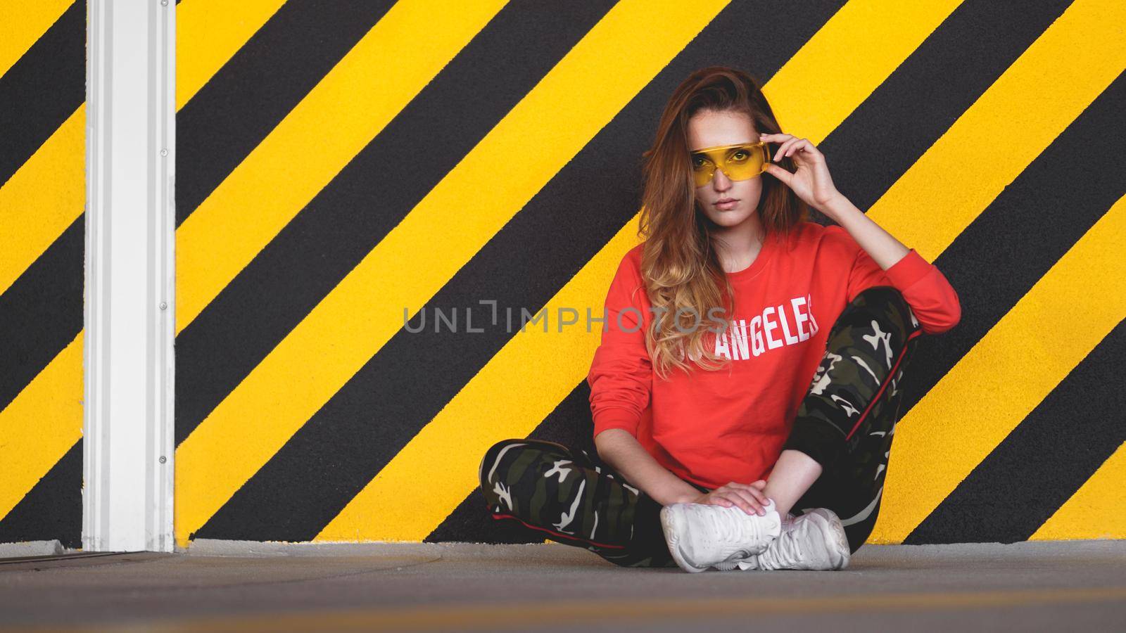 Fashion model wearing red hoodie with the inscription los angeles and big plastic glasses posing in the city at parking. Fashion urban outfit